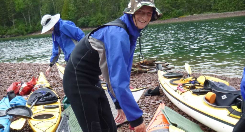 A person smiles at the camera while tending to kayaks resting on the shore of a lake. A person does the same in the background. 